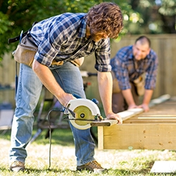 Mid adult carpenter cutting wood with handheld saw while coworker helping him in background at construction site